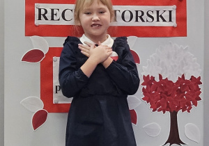 Uczennica w galowym stroju na tle ozdobnej tablicy z napisem Konkurs recytatorski - wiersze patriotyczne.