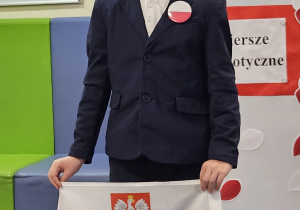 Uczeń w galowym stroju na tle ozdobnej tablicy z napisem Konkurs recytatorski - wiersze patriotyczne.