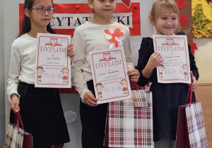 Trzy uczennice w strojach galowych z dyplomami i torbami upominkowymi w dłoniach na tle ozdobnej tablicy z napisem Konkurs recytatorski - wiersze patriotyczne.