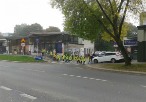 Grupa uczniów w kamizelkach odblaskowych, idąca parami, w kolumnie, przechodząca za nauczycielką przez przejście dla pieszych.
