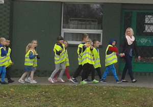 Nauczycielka idąca z pierwszą parą będąca na czele kolumny uczniów w kamizelkach odblaskowych, idących parami po chodniku.
