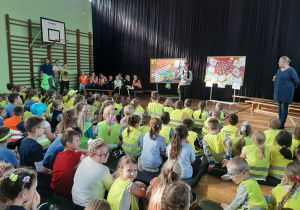 . Uczniowie siedzą na materacach ubraniu w odblaskowe kamizelki, przed nimi stoi nauczycielka.