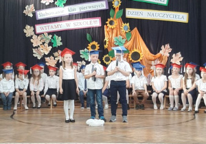 Troje uczniów stoi na środki sali, jeden z uczniów trzyma w ręku mikrofon i mówi wiersz
