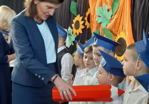 Pani Dyrektor pasuje czerwoną kredką uczniów w niebieskich biretach.