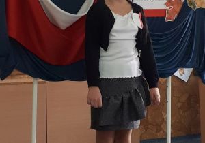 Uczennica w stroju galowym stoi na tle udekorowanej tablicy z napisem: Konkurs recytatorski. Wiersze patriotyczne.