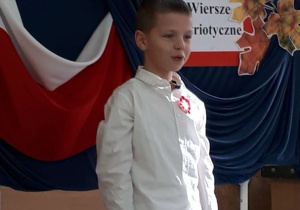 Uczeń w stroju galowym stoi na scenie i recytuje wiersz.