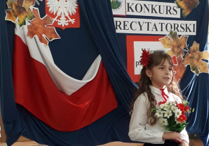 Uczennica w stroju galowym, z bukietem kwiatów stoi na tle udekorowanej tablicy.