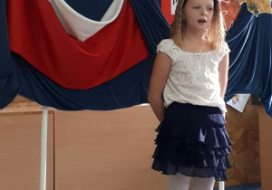 Uczennica w stroju galowym stoi na scenie i mówi wiersz.