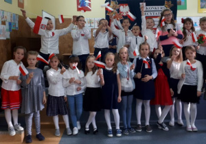 23 osobowa grupa uczniów z klas I-III, w odświętnych strojach, z chorągiewkami w dłoniach stoi na tle udekorowanej tablicy z napisem: Konkurs recytatorski. Wiersze patriotyczne.