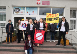 Grupa uczniów trzymających transparenty stoi wraz z koordynatorem projektu przed budynkiem szkoły.