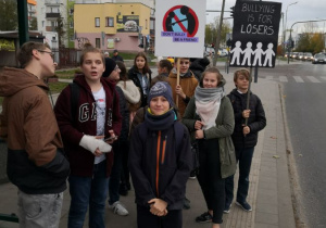 Grupa uczniów trzymających transparenty stoi na przystanku autobusowym.