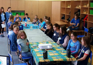 Uczniowie w niebieskich strojach (kolor Unicefu) z logo Międzynarodowego Dnia Dziecka siedzą przy stole dyskusyjnym.