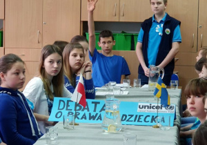 Uczeń z klasy starszej stoi przy stole dyskusyjnym i odpowiada na pytania, uczeń siedzący obok niego ponosi rękę.