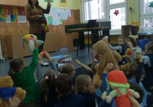 Pani Lektorka stoi na scenie i ma na ręku Pacynkę Misia, uczniowie siedzą na podłodze i podnoszą do góry swoje Misie.