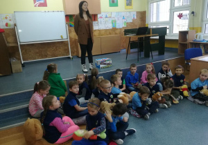 Pani Lektorka stoi na scenie, a uczniowie siedzą pod sceną z uwagą słuchając opowieści o misiu Teddy Eddi.