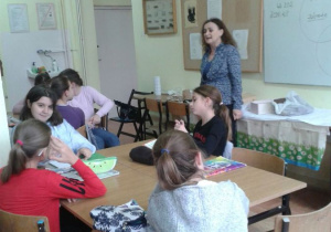 Dzieci siedzą przy stołach, nauczyciel opowiada o sposobie wykonania zadania.