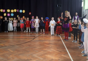Liczna grupa dzieci i dwoje dorosłych w przebraniach stoi w kole na udekorowanej sali gimnastycznej, mają uniesione ręce.