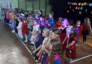 Liczna grupa dziewczynek w przebraniach stoi wzdłuż białej linii, skierowana twarzą w lewą stronę.