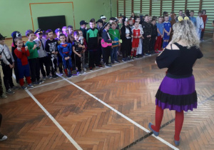 Liczna grupa chłopców w przebraniach stoi wzdłuż białej linii, na wprost nauczycielki w przebraniu.