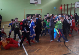 Z lewej strony stoją dwie nauczycielki w przebraniach, pośrodku liczna grupa chłopców w przebraniach jest w ruchu (tańczy).
