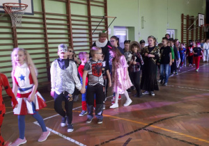 Liczna grupy dzieci i dwie nauczycielki będący w przebraniach tworzą korowód na dużej sali.