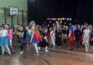Liczna grupa dzieci w przebraniach stoi w rozsypce na udekorowanej sali gimnastycznej.