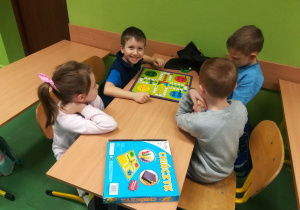 Trzech chłopców i jedna dziewczynka grają w "Chinczyka", jeden z chłopców uśmiecha się do nauczyciela.