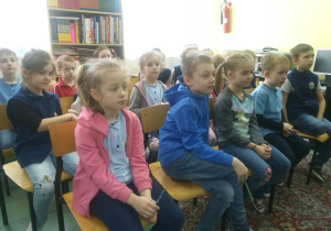 Uczniowie siedzą na krzesłach i słuchają powitania nauczycielki.