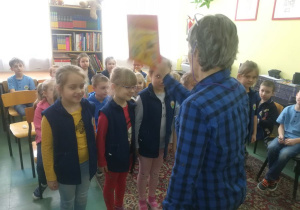 Nauczycielka pasuje uczennicę na czytelniczkę dotykając jej ramienia książką.