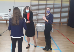 Nauczycielka wręcza nagrodę uczennicy, obok stoi druga nauczycielka, w tle widać 3 inne uczennice, ubrane na galowo, zdjęcie jest wykonane na sali gimnastycznej, za nauczycielką stoi stolik.