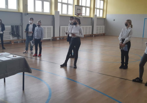 Na zdjęciu widać salę gimnastyczną, 2 uczniów i 6 uczennic stoi w dwóch rzędach w odstępach, są ubrani na galowo, trzymają w rękach nagrody, między nimi stoi jedna nauczycielka, za nimi druga.