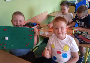 Czterech chłopców siedzących w ławkach. Jeden z nich trzyma pracę plastyczną w kropki