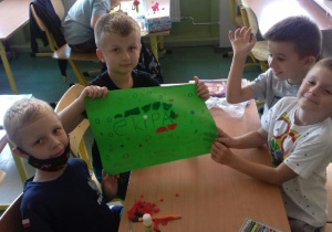 Czterech chłopców siedzących przy jednej ławce. Trzymają pracę plastyczną w z napisem "ekipa", z narysowanymi dookoła kropkami