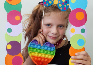 Uśmiechnięta dziewczynka, trzymająca kolorową ramkę, pop it w kształcie serca i kokardę w kropki nad głową