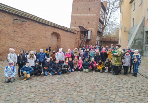 Grupa dzieci wraz z panem przewodnikiem ubranym w zielony płaszcz, pozuje do zdjęcia na tle starych murów miasta