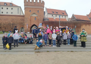 Grupa dzieci wraz z panem przewodnikiem ubranym w zielony płaszcz, pozuje do zdjęcia na tle Bramy do Starego Miasta
