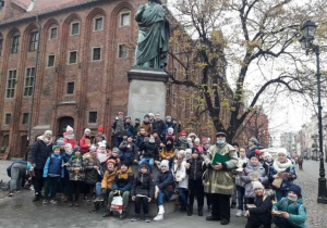 Grupa dzieci wraz z panem przewodnikiem ubranym w zielony płaszcz, pozuje do zdjęcia pod pomnikiem Mikołaja Kopernika