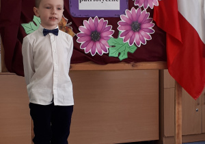 Uczeń w galowym stroju na tle ozdobnej tablicy z napisem Konkurs recytatorski - wiersze patriotyczne.