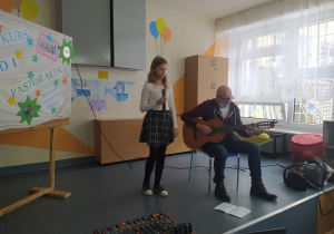 Scena, na której stoi śpiewająca dziewczynka, a obok niej siedzi na krześle jej tata, który akompaniuje jej na gitarze.