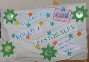 Korkowa tablica na której wykonany jest napis "Konkurs Kolęd i Pastorałek, Śpierwajmy i grajmy Mu, Małemu" oraz są przyczepione ozdoby tj. zielono - srebne gwiazdy, świąteczne obrazki.