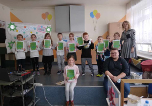 Grupa dzieci z klas I - III stoi na scenie z dyplomami w ręku. Obok nich stoi nauczycielka, a zarazem prowadząca konkurs. Na brzegu sceny siedzi dziewczynka z dyplomem, a obok niej siedzi nauczyciel muzyki, członek Jury.