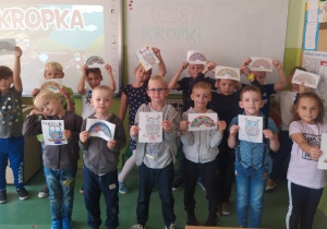 Grupa dzieci trzymająca prace plastyczne wykonane z kropek na tle tablicy z napisem Dzień Kropki