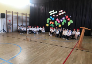 Pierwszoklasiści siedzący na ławkach z galowych strojach w biretach na głowach