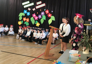 Pierwszoklasiści siedzą na ławkach a uczniowie klas starszych przemawiają do zgromadzonych gości