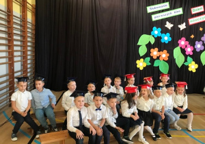 Pierwszoklasiści siedzą na ławkach w galowych strojach w biretach na głowach