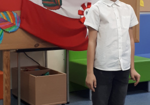 Uczeń w stroju galowym na tle tablicy z napisem "Konkurs recytatorski - wiersze patriotyczne"