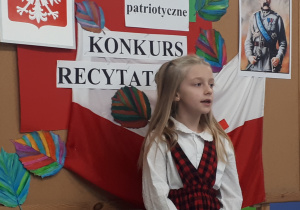 Uczennica w stroju galowym recytująca wiersz na tle ozdobnej tablicy z napisem "Konkurs recytatorski - wiersze patriotyczne"