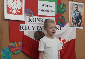 Uczeń w stroju galowym na tle ozdobnej tablicy z napisem "Konkurs recytatorski - wiersze patriotyczne"