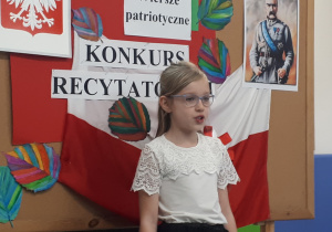 Uczennica w stroju galowym na tle ozdobnej tablicy z napisem "Konkurs recytatorski - wiersze patriotyczne"