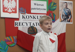 Uczeń w stroju galowym na tle tablicy "Konkurs recytatorski - wiersze patriotyczne"